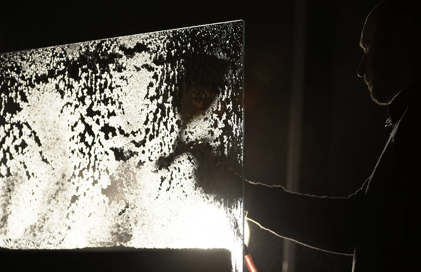 A large sheet of glass covered in frost, lit from behind so the ice crystals glow white against black, with a technician silhouetted beside it, cable trailing to a light rig.