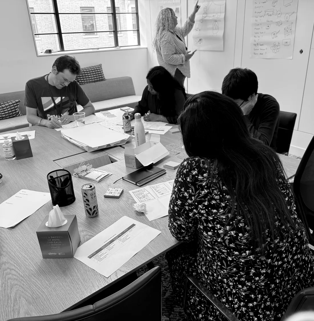 A cross-functional workshop in progress: participants around a table covered in printed journey screens and worksheets, with a facilitator annotating a whiteboard.